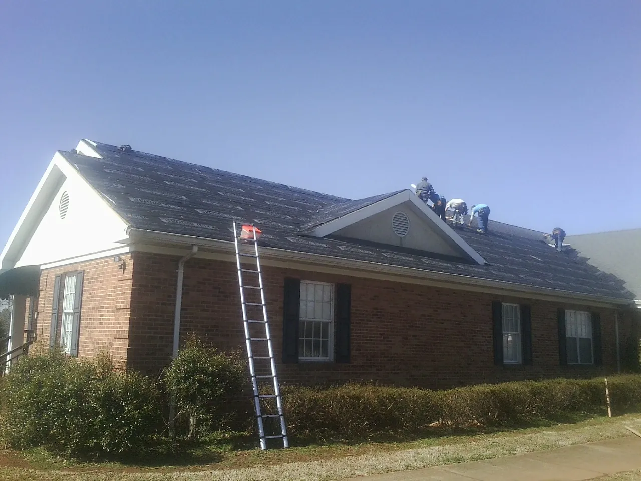 Crew tearing shingles off a brick ranch roof with a ladder set — Metal Roof Repair work by our crew in Youngsville